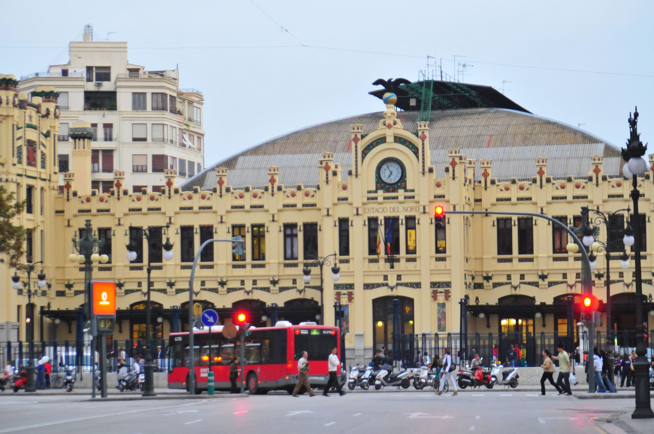 Historisches Bahnhofsgebäude mit Uhr, rotem Stadtbus und Fußgängern an einer Straße. Historisches Bahnhofsgebäude mit Uhr, rotem Stadtbus und Fußgängern an einer Straße.
