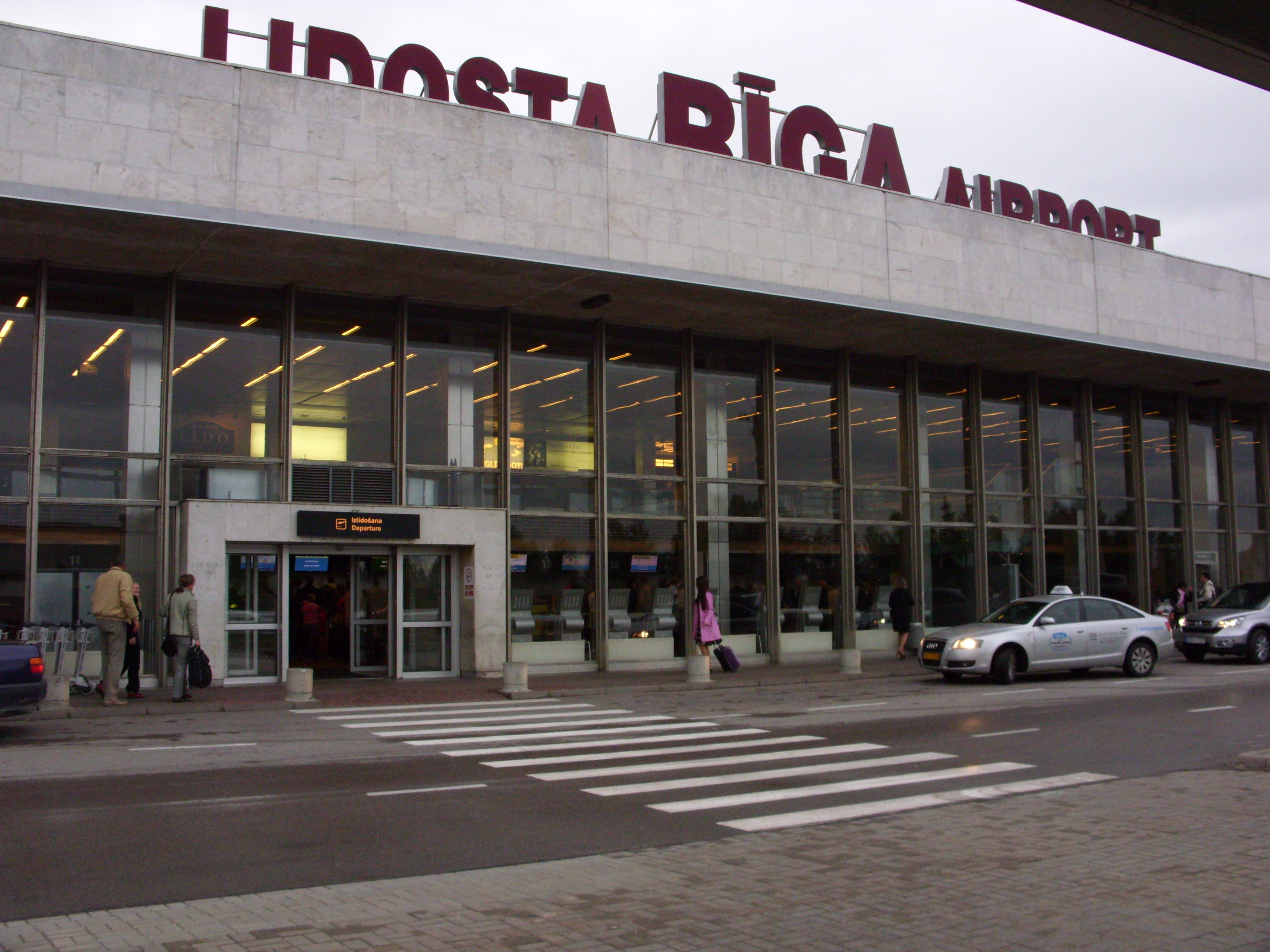 Außenansicht des Flughafengebäudes in Riga mit Eingang und Reisenden mit Gepäck.
